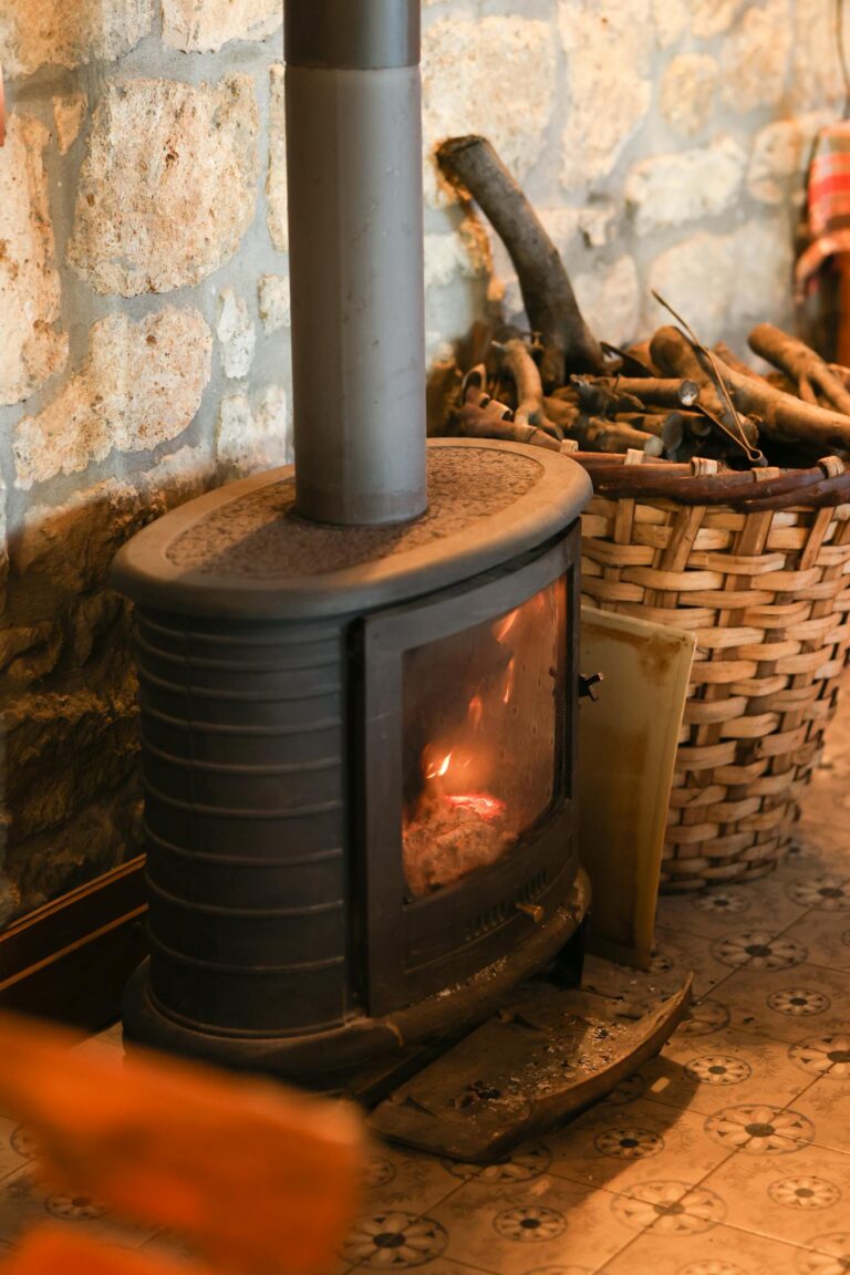 Warm and rustic indoor scene featuring a lit wood stove by a stone wall with a basket of firewood.